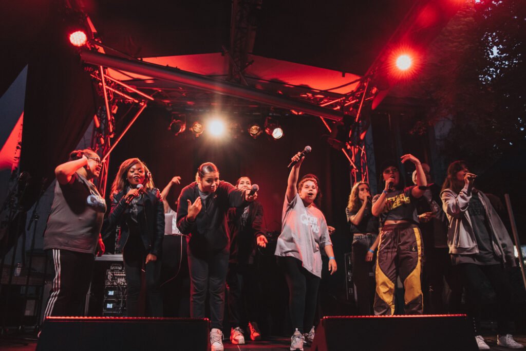 Eine Gruppe von weiblich gelesenen Schwarz, of Color und weißen Menschen steht in zwei Reihen auf einer Bühne. Personen im Vordergrund halten Mikrofone und rappen.