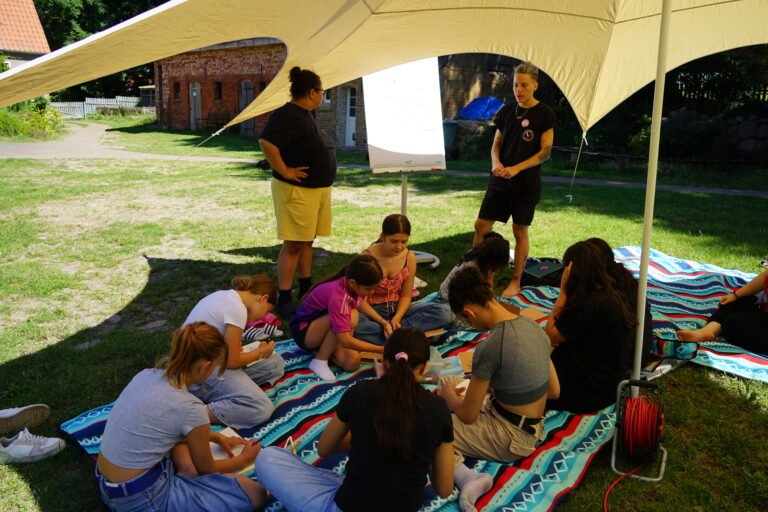 Mehrere Jugendliche sitzen auf einer Decke unter einem Pavillon und schreiben. Hinter ihnen stehen zwei weitere Personen, die etwas an einem Flip-Chart erklären