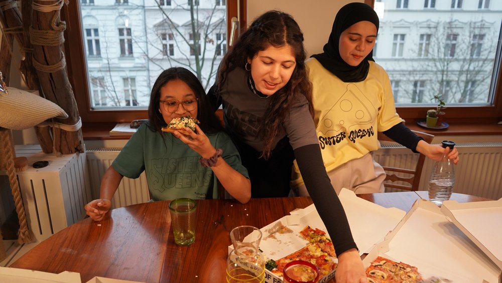 Drei weiblich gelesene Personen sitzen und stehen hinter einem Tisch und greifen nach Pizzastücken aus einer Schachtel