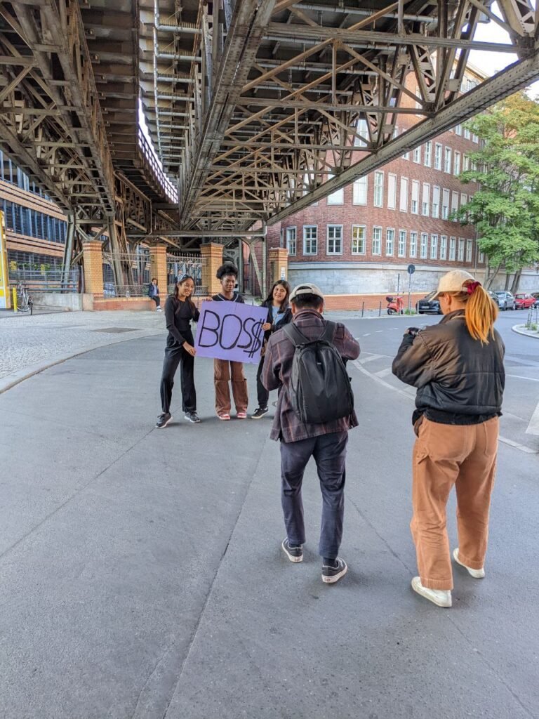 Drei Mädchen of Color stehen unter einer Brücke und halten ein Schild mit der Aufschrift „BOSS“ hoch. Vor ihnen stehen zwei Personen, die sie filmen.