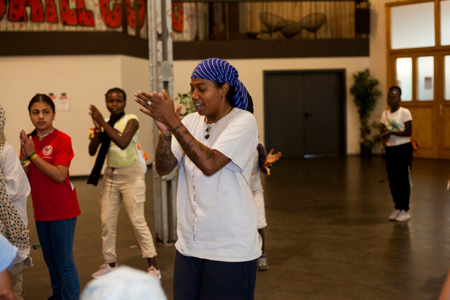 Eine Schwarze Frau steht in einer Halle und tanzt. Um sie herum stehen Jugendliche und imitieren ihre Bewegungen