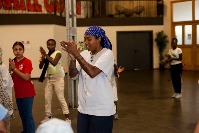 Eine Schwarze Frau steht in einer Halle und tanzt. Um sie herum stehen Jugendliche und imitieren ihre Bewegungen