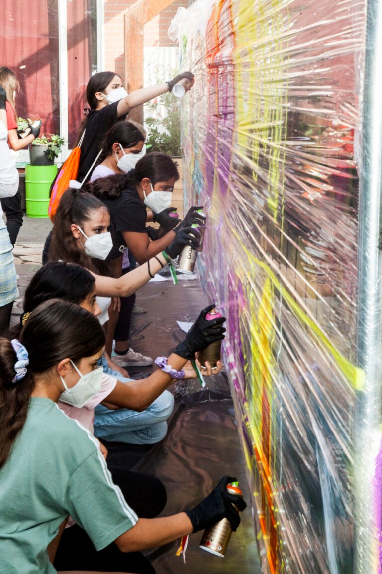 Mehrere Jugendliche stehen vor einem mit Folie bespannten Bauzaun und besprühen diesen mit Graffitis