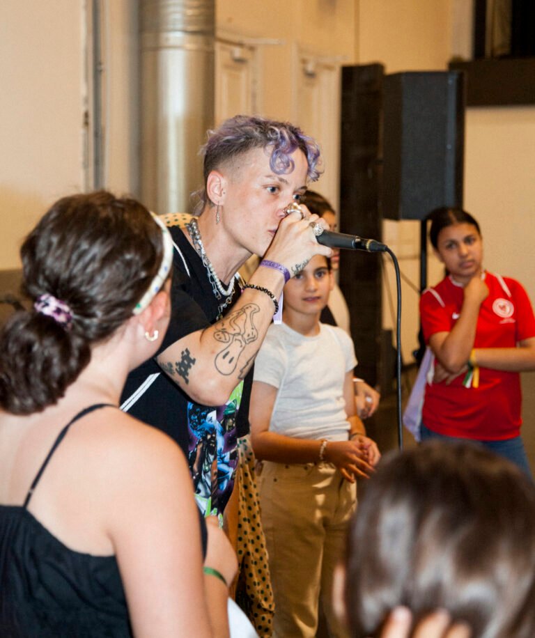 Eine Person rappt in ein Mikrofon. Um die Person stehen mehrere Jugendliche und schauen zu