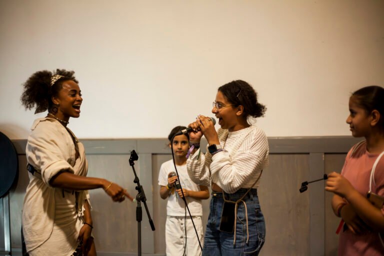 Zwei Frauen of color stehen sich gegenüber und rappen. Im Hintergrund und am rechten Rand steht ieweils ein Mädchen