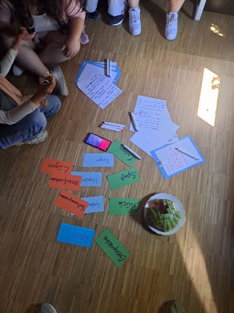 Viele Zettel und Stifte liegen auf dem Fußboden verteilt. Am oberen Bildrand sitzen Personen. Es sind nur die Füße auf dem Bild dargestellt.