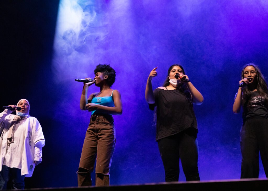 Vier weiblich gelesene Jugendliche of Color stehen auf einer Bühne und rappen.