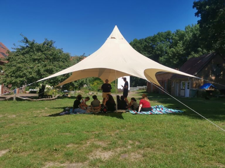 Eine Gruppe von Menschen sitzt in einem Garten unter einem Pavillion. Der Pavillion ist rund und läuft nach oben spitz zu.